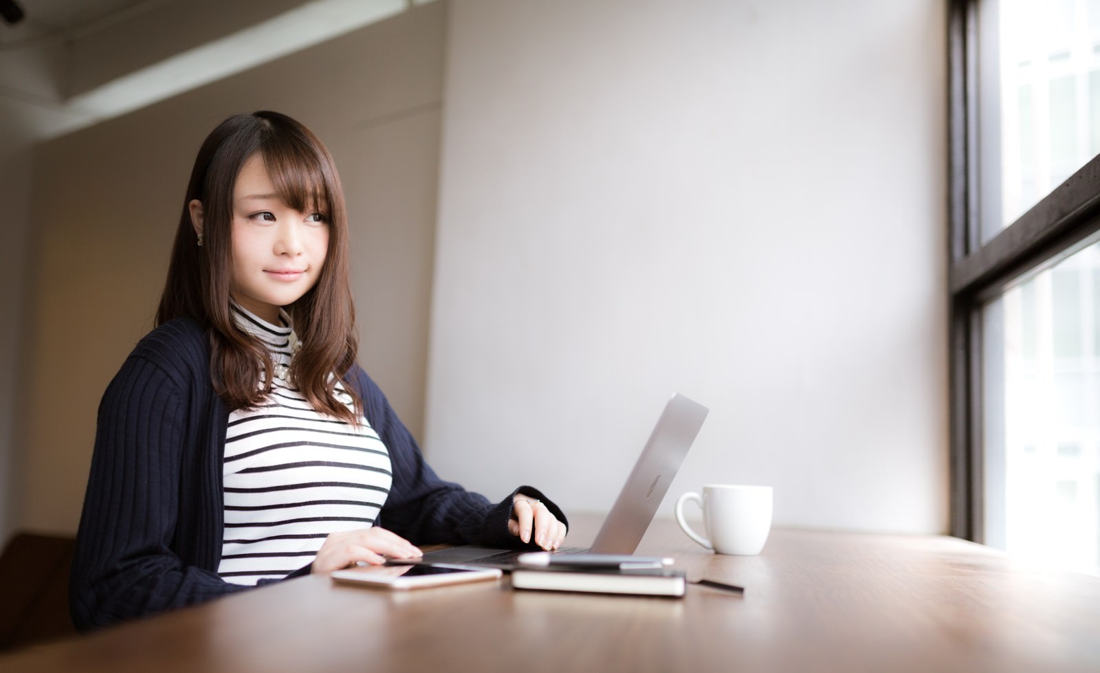 カフェでノートパソコンを使って作業する女性の横向き写真［モデル：茜さや］