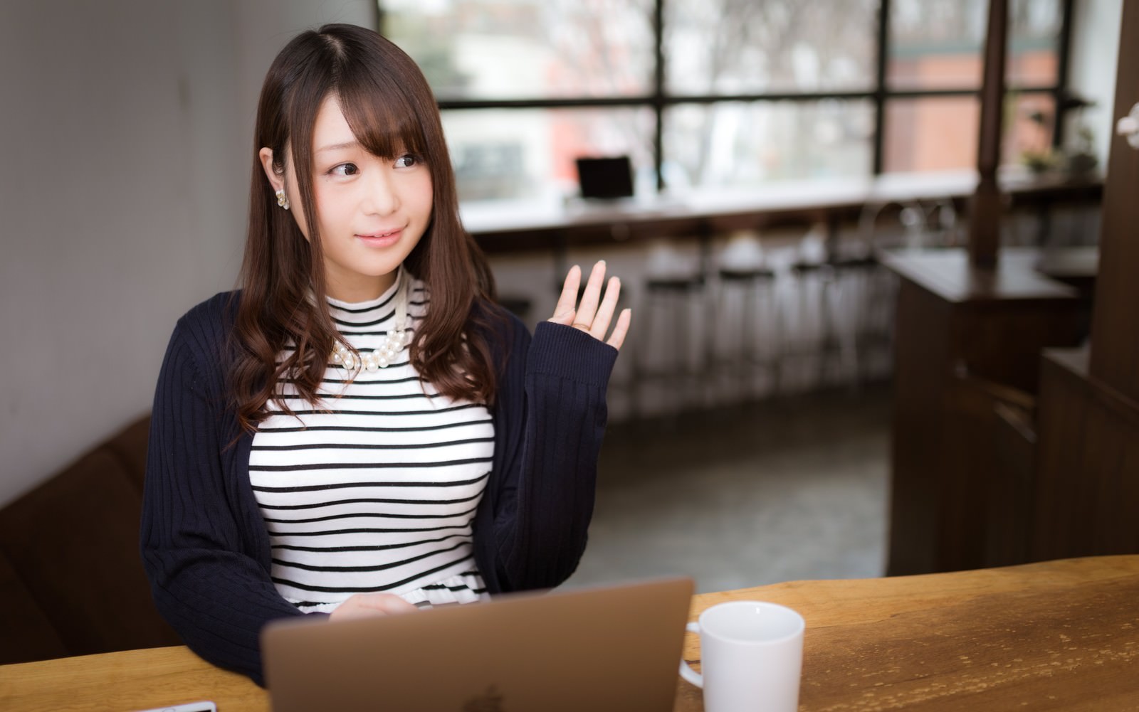カフェでラップトップを前に挨拶をする女性［モデル：茜さや］