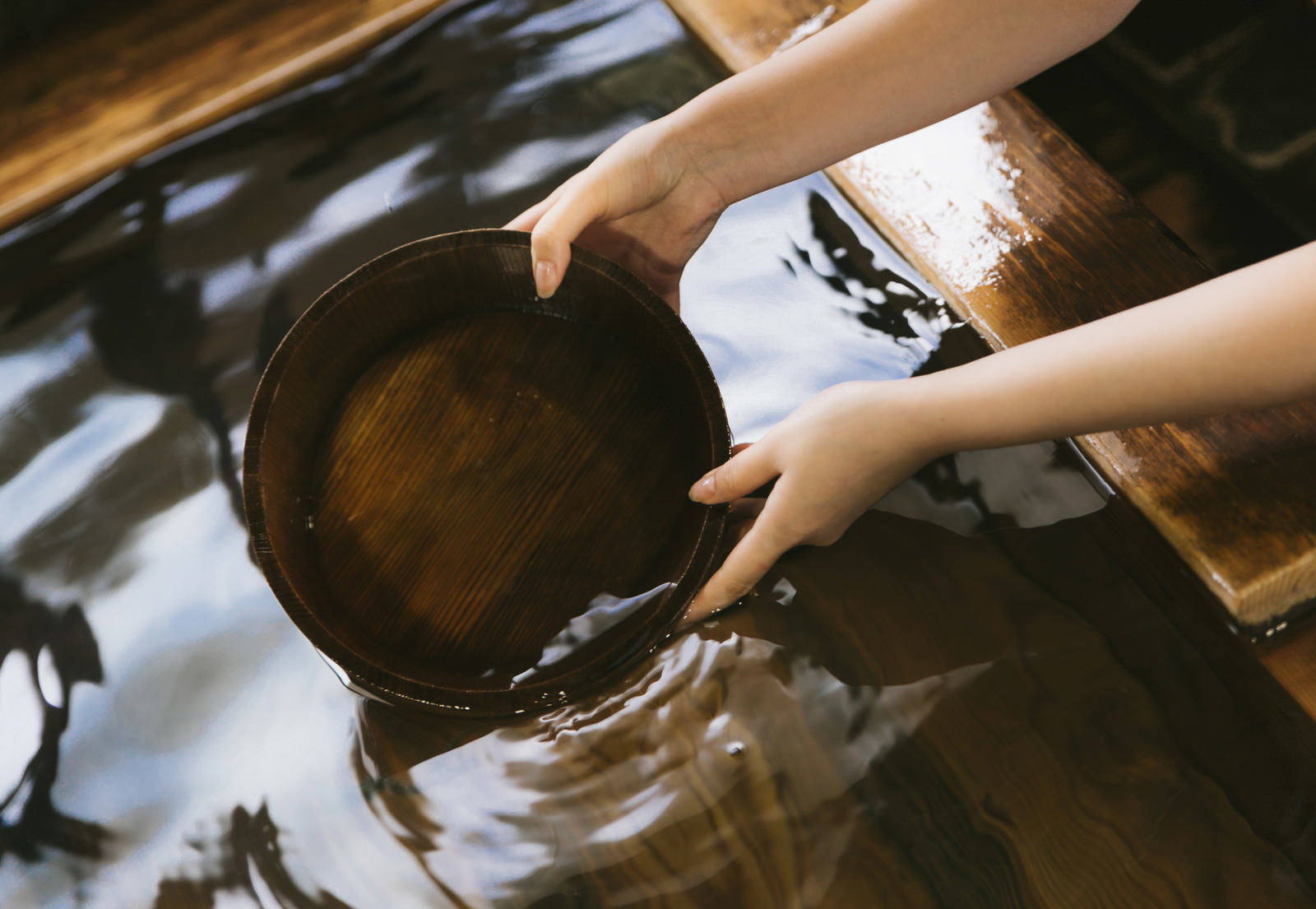 温泉の檜風呂で湯船にかけ湯の桶を入れる女性の手元
