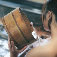 檜風呂で桶を使って肩にかけ湯する女性の後ろ姿の写真