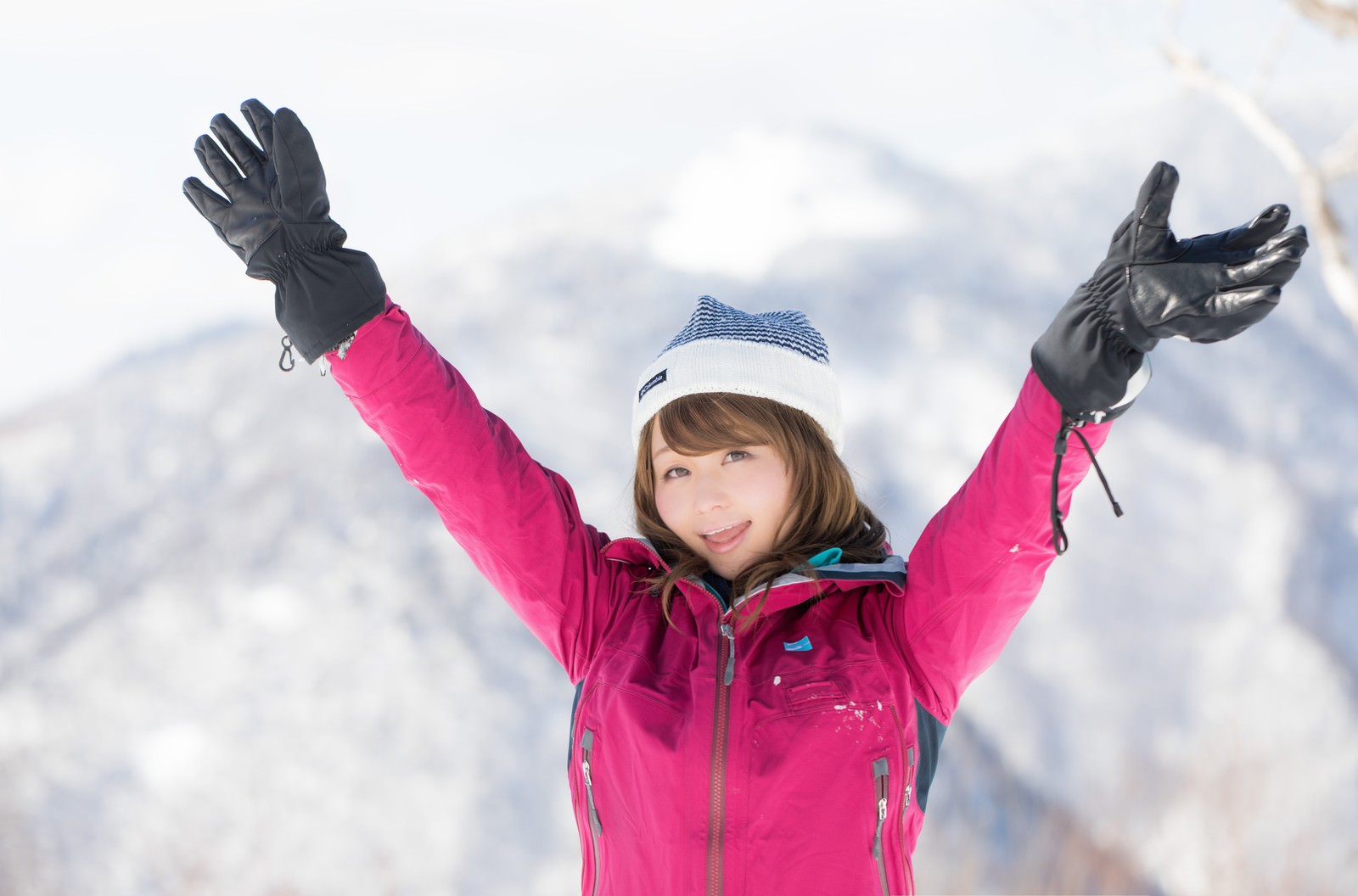 ピンクのウェアを着た女性が雪の山頂で両腕を広げて上を向いている［モデル：茜さや］