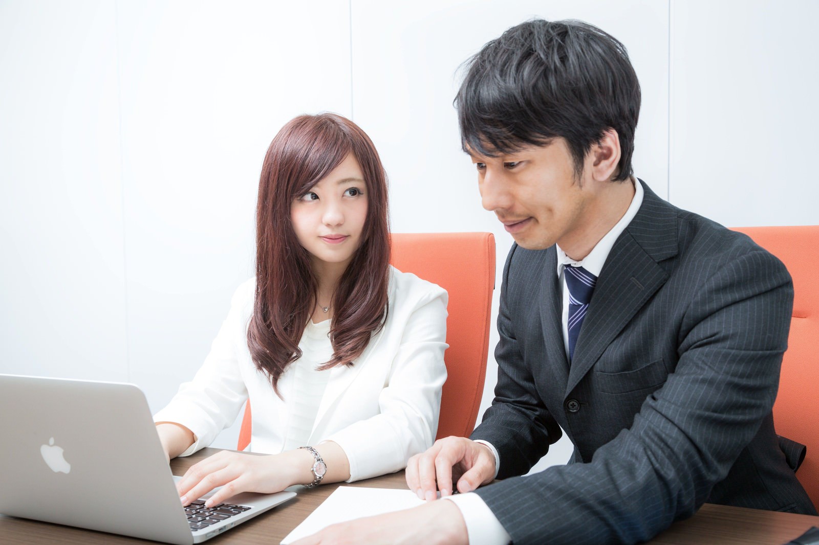 オレンジのソファーに座り、男性を見つめる白いジャケット姿の新入社員女性［モデル：大川竜弥 河村友歌］