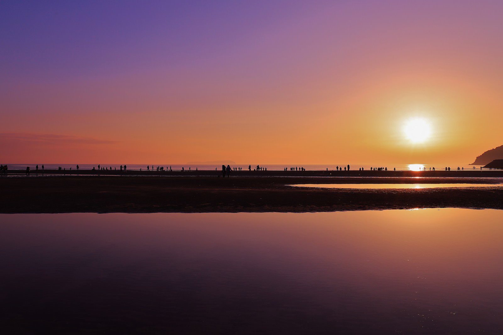 夕焼け空の下、浜辺で遊ぶ人々のシルエットと海面に映る夕日
