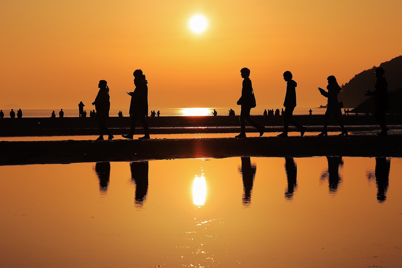 夕暮れ時の砂浜で散策する人々のシルエットと水面に映る夕日