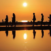 夕暮れ時に砂浜を散策する人達のシルエットと夕日のリフレクションの写真