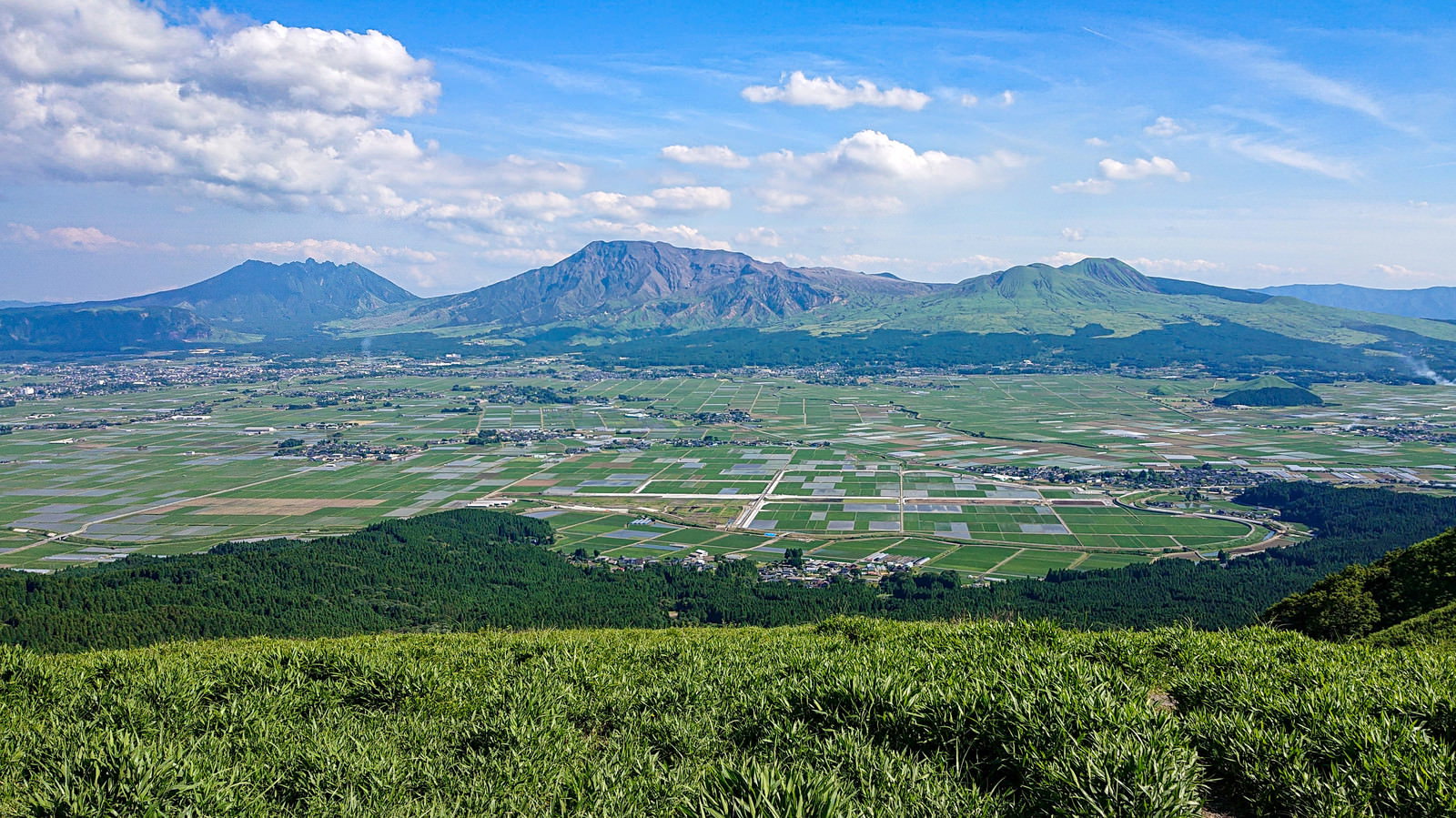 青空に雲が浮かぶ中、緑の山脈と田園風景が広がるパノラマ風景