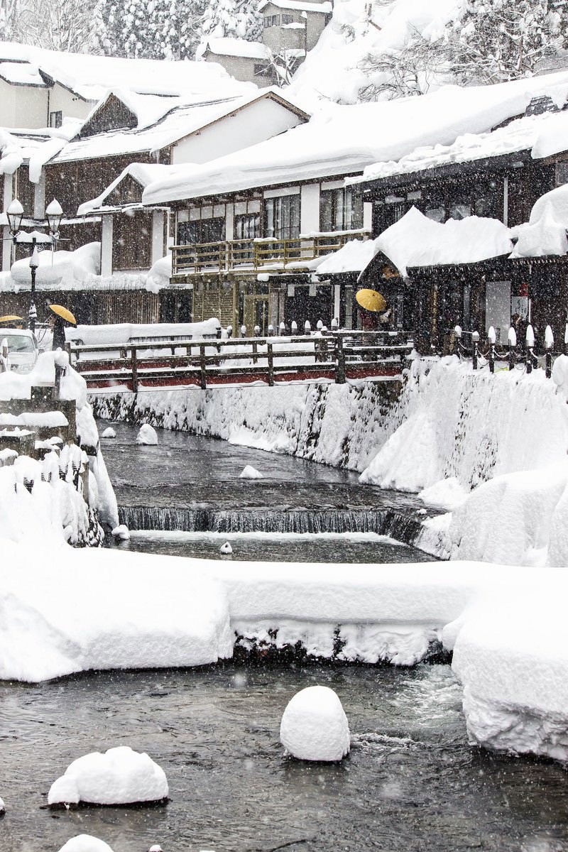 雪が積もる銀山温泉街で、旅館が両側に立ち並び、川沿いの橋を歩く観光客の風景
