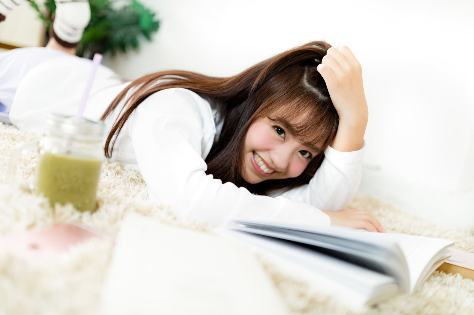グリーンスムージーを持ちながらラグの上で読書する笑顔の女性［モデル：河村友歌］