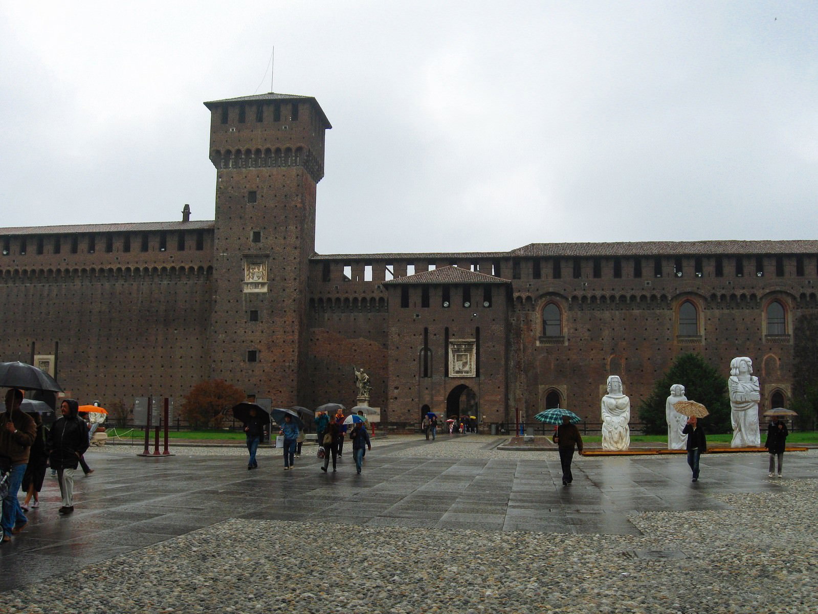 イタリア・ミラノのスフォルツェスコ城前の石畳広場で傘を差して歩く観光客たち