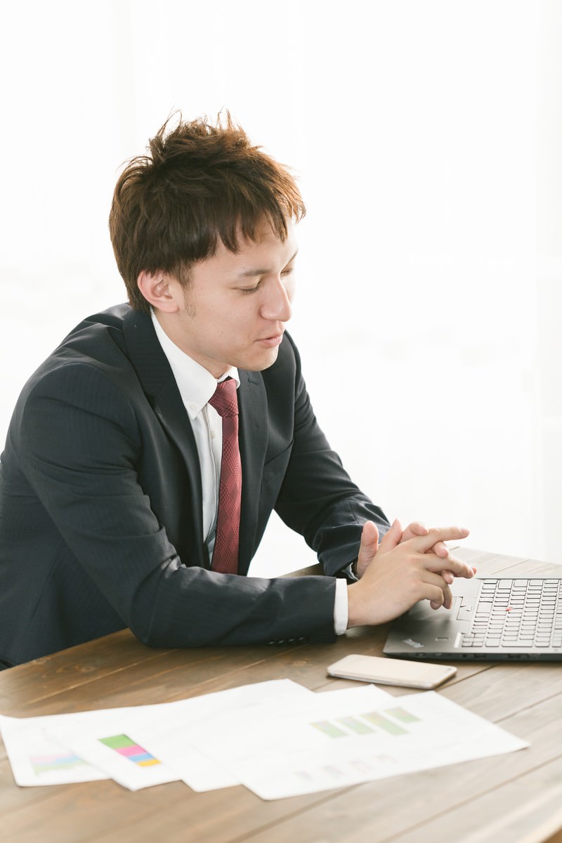 机に向かい資料を見ながらプランを提示するスーツ姿の営業マン［モデル：鈴木秀］