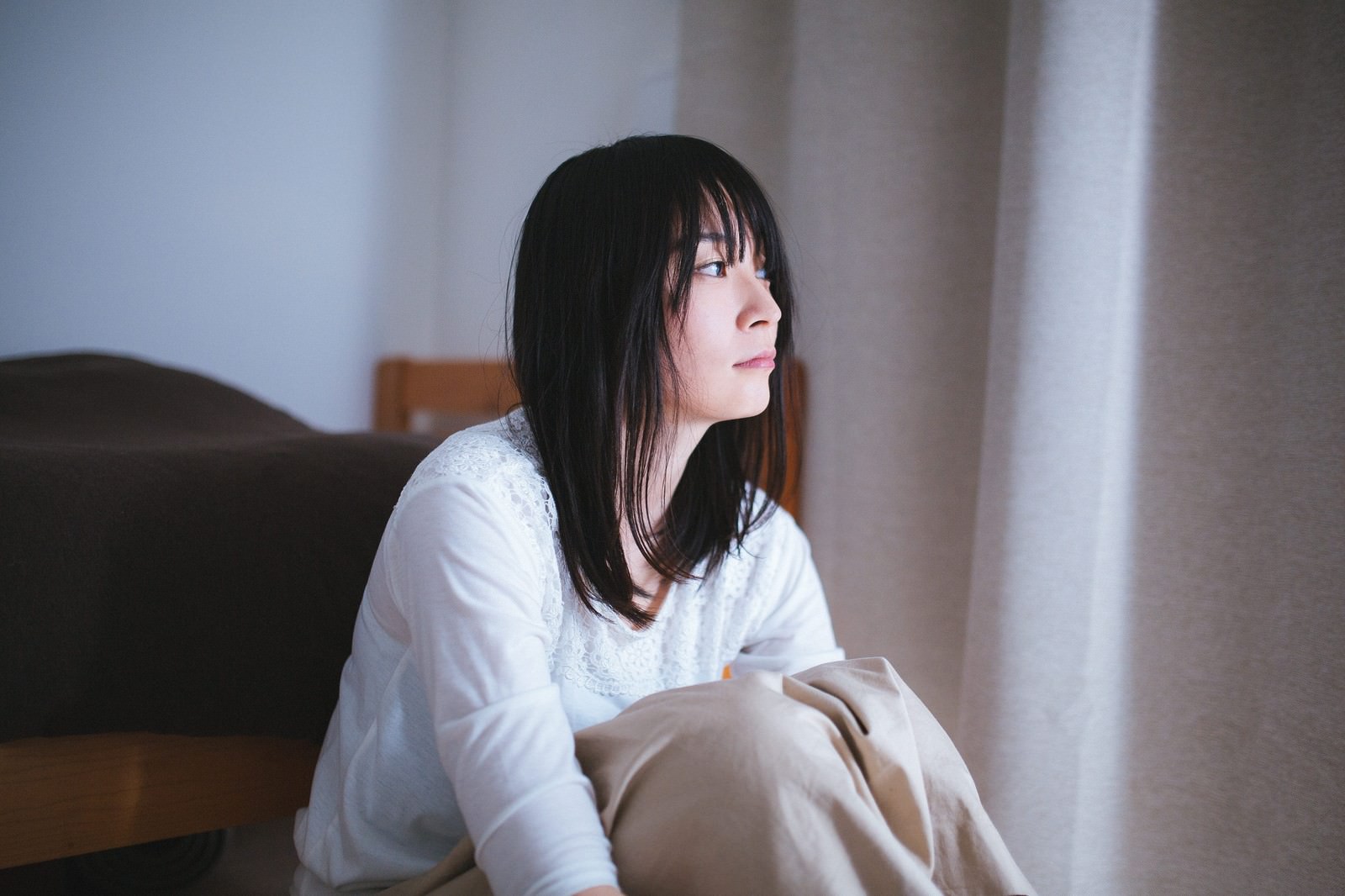 ソファーに座ってうつむく白いシャツの女性。朝日が窓から差し込む室内［モデル：たけべともこ］