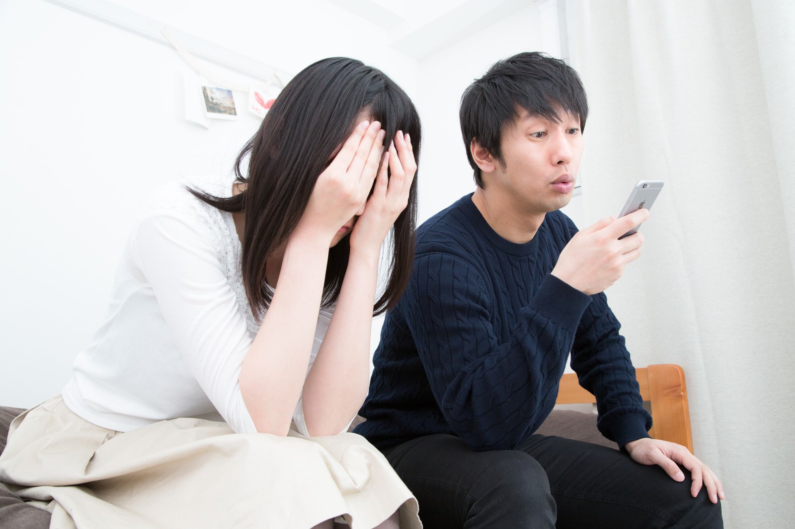 泣く女性の隣でスマートフォンを見つめる男性。室内で向き合うカップルの対比的な表情［モデル：大川竜弥 たけべともこ］