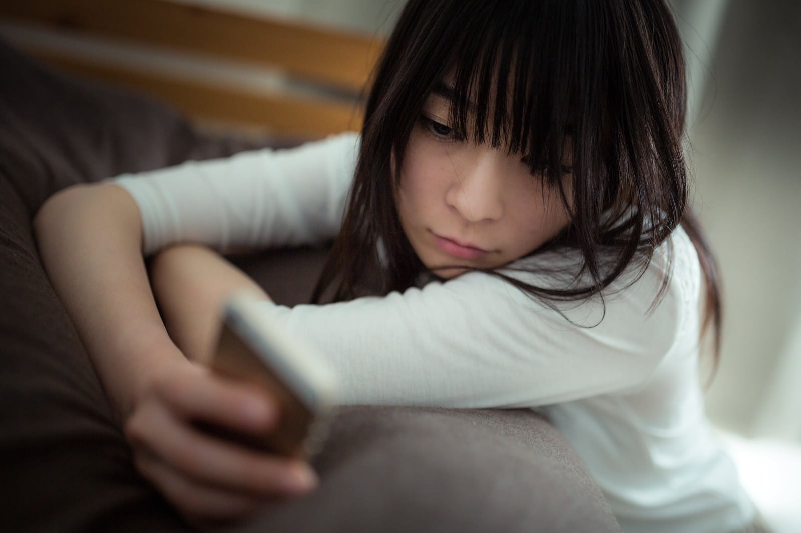 ソファーに座ってスマホを見つめる失恋した女性［モデル：たけべともこ］