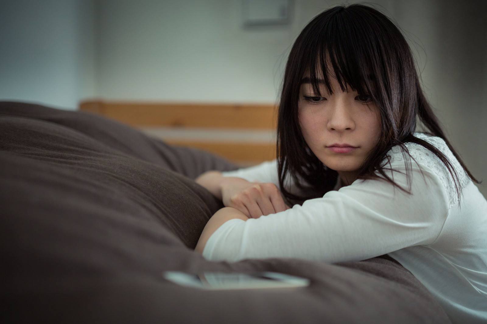ベッドに寝転びスマホを握って画面を見つめる女性［モデル：たけべともこ］