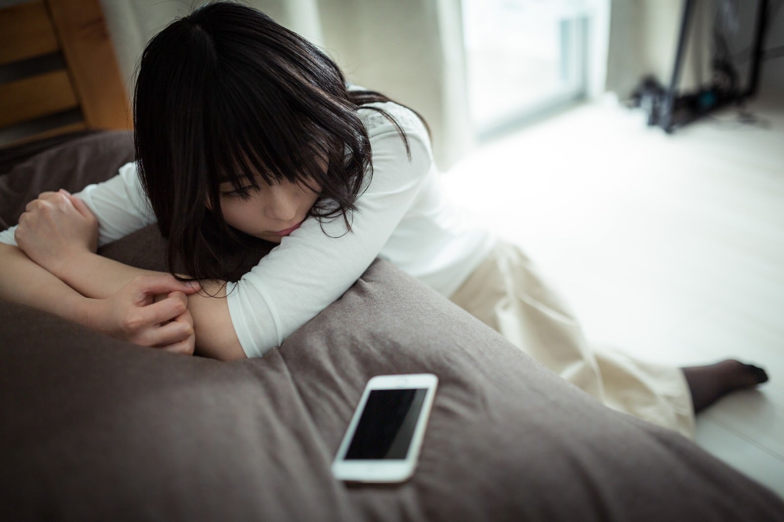 床に座り、スマートフォンを前に下を向いている失恋した女性［モデル：たけべともこ］