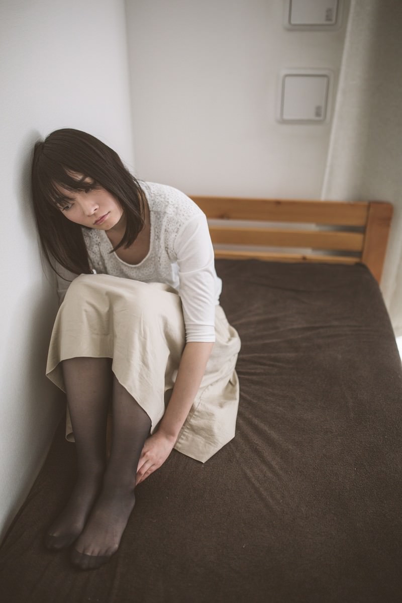 ベッドの上で膝を抱えて下を向く女性［モデル：たけべともこ］