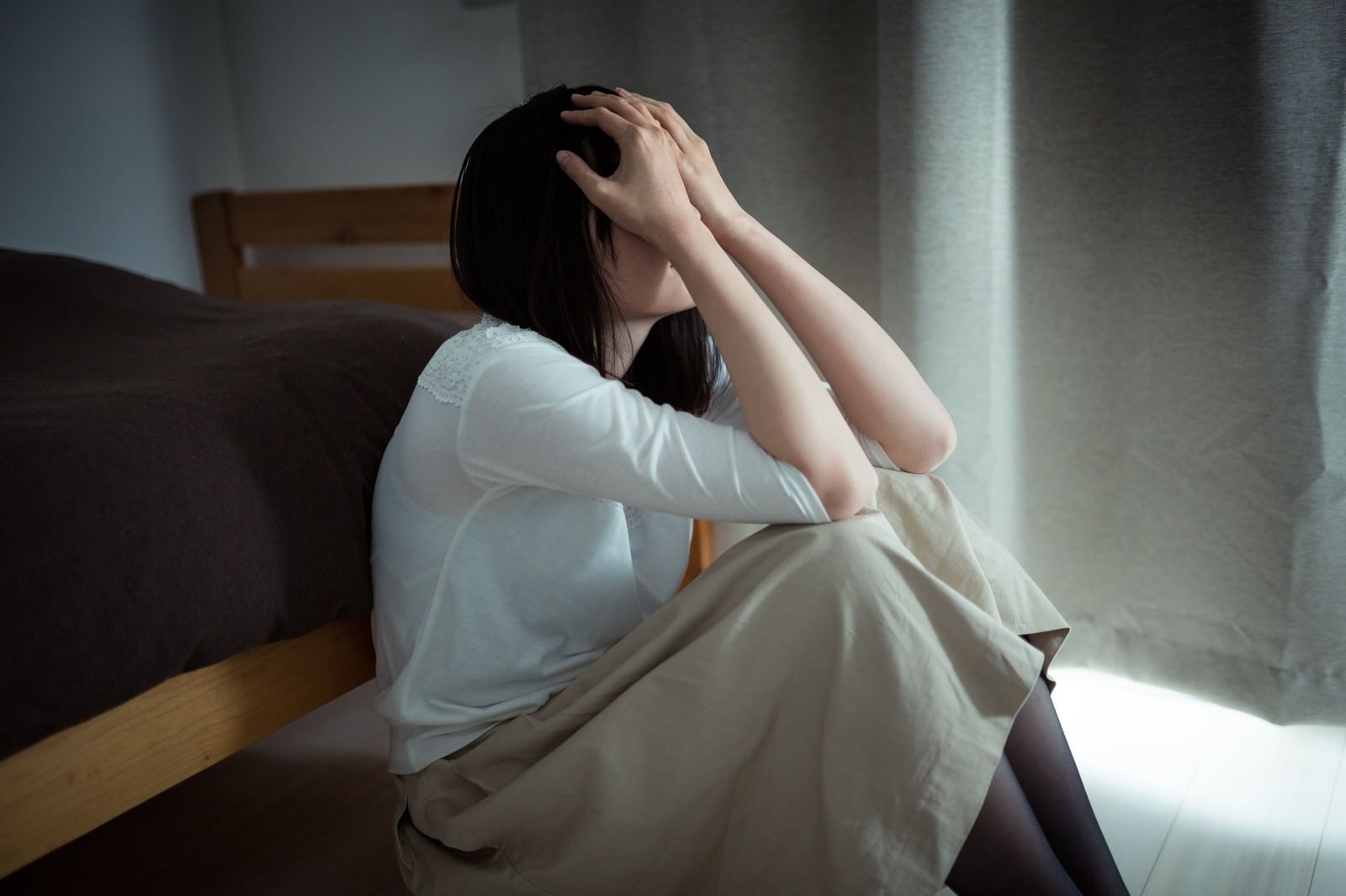 白いTシャツを着た女性が椅子に座り、両手で顔を覆いながら泣いている［モデル：たけべともこ］