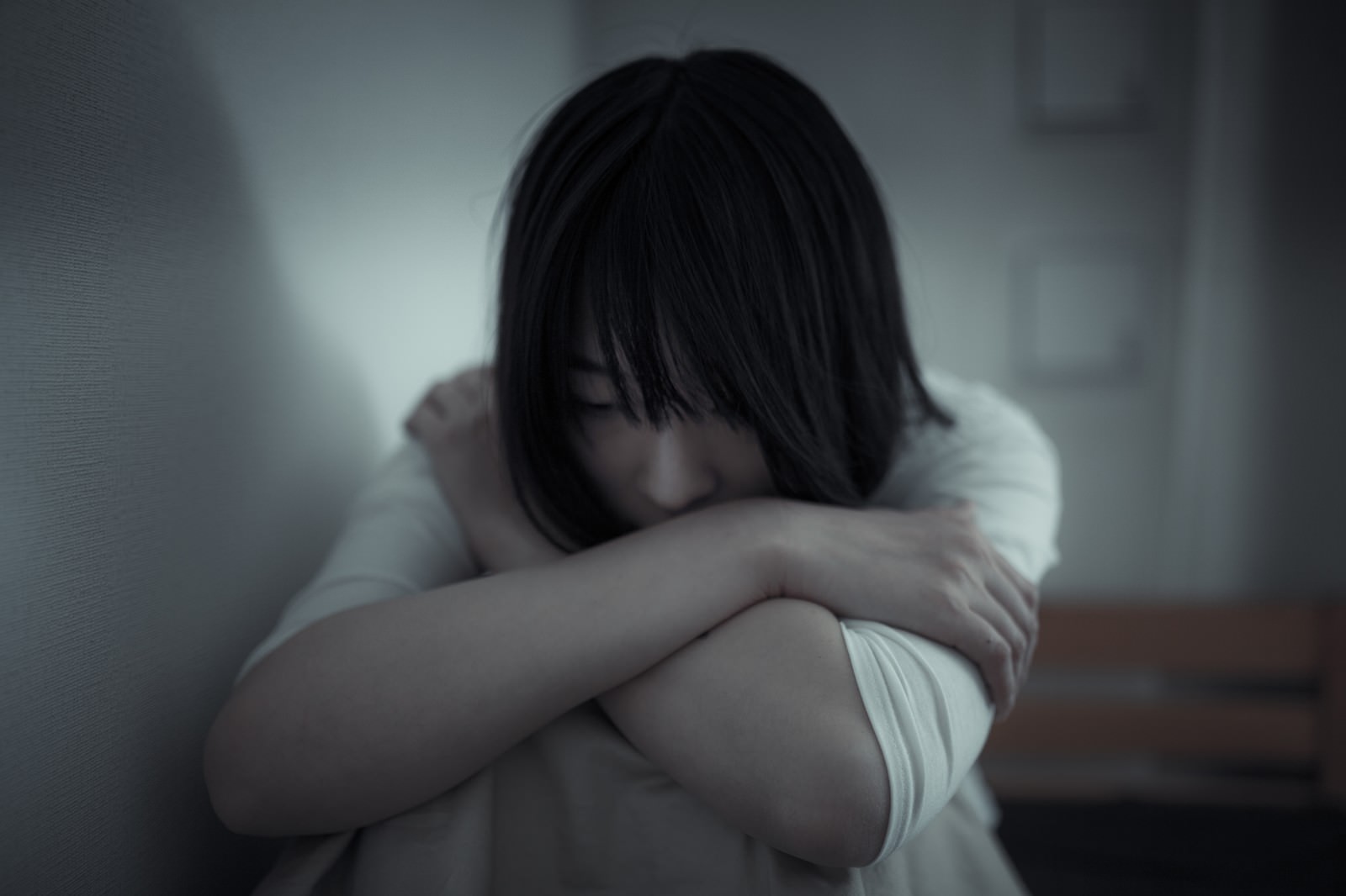 絶望した表情で体育座りをする黒髪の女性。白いトップスを着て下を向いている［モデル：たけべともこ］