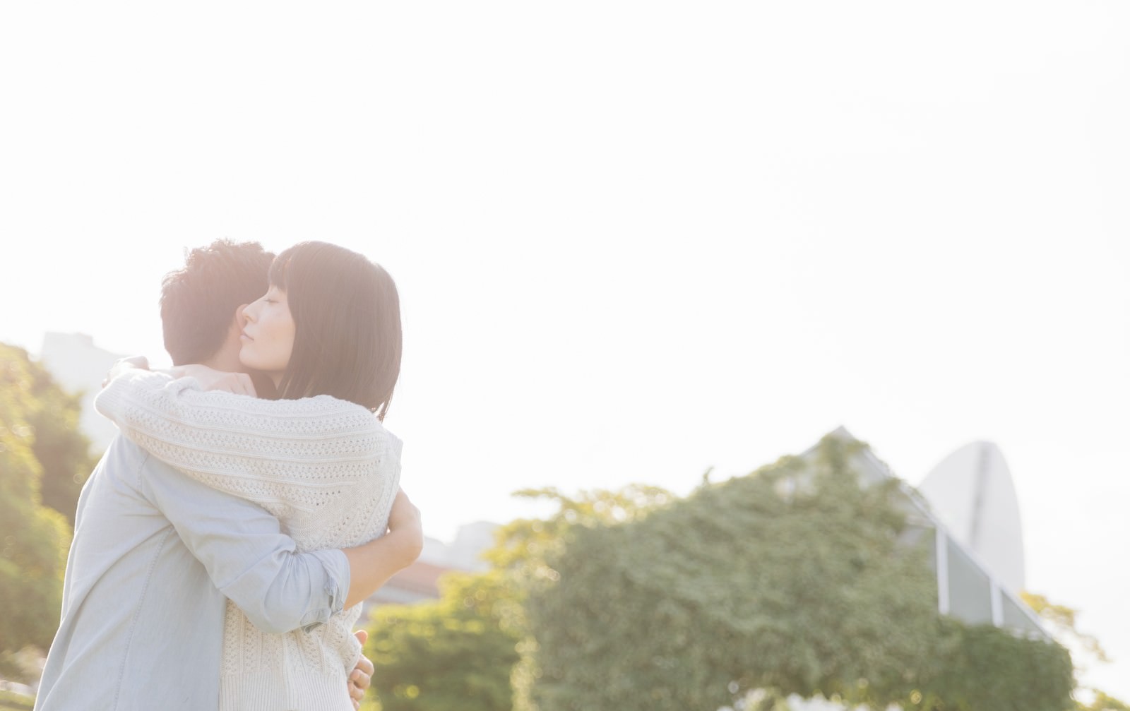 逆光の中で男性が女性を優しく抱きしめているカップルのハグシーン［モデル：たけべともこ］