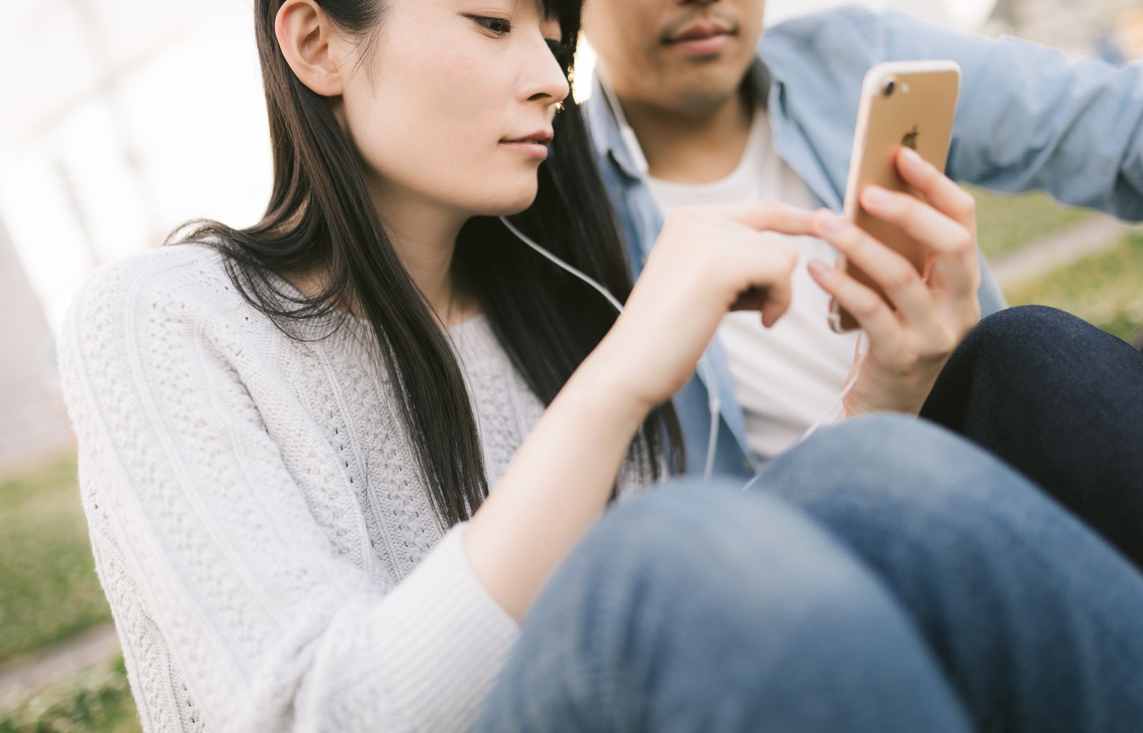 白いニットを着た女性がイヤホンをしながら彼氏とスマホを見ている［モデル：たけべともこ］