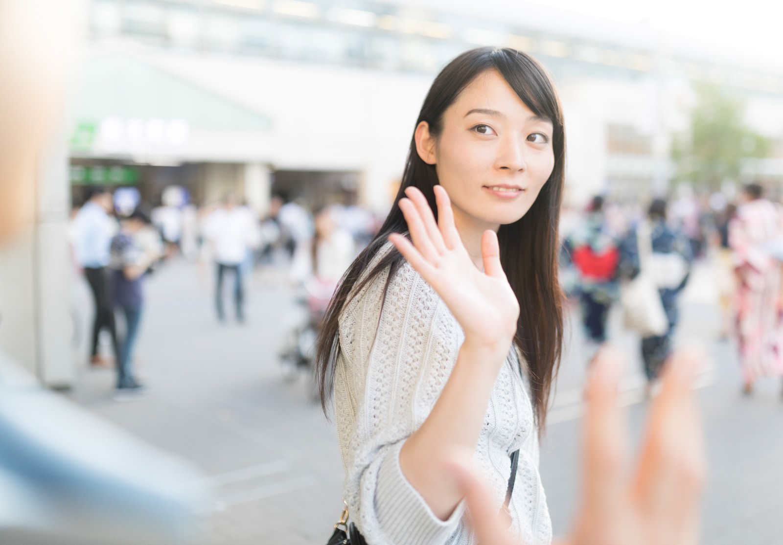 駅前で振り返りながら手を振る白いトップスの女性［モデル：たけべともこ］