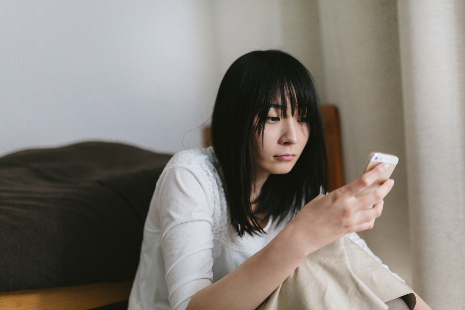 ソファーに座ってスマートフォン画面を見つめる黒髪の女性［モデル：たけべともこ］