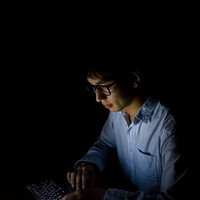 暗い部屋でノートPCの画面に照らされる男性の写真