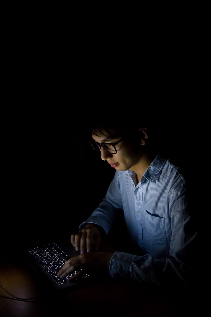 暗い部屋でPCに照らされる男性［モデル：大川竜弥］