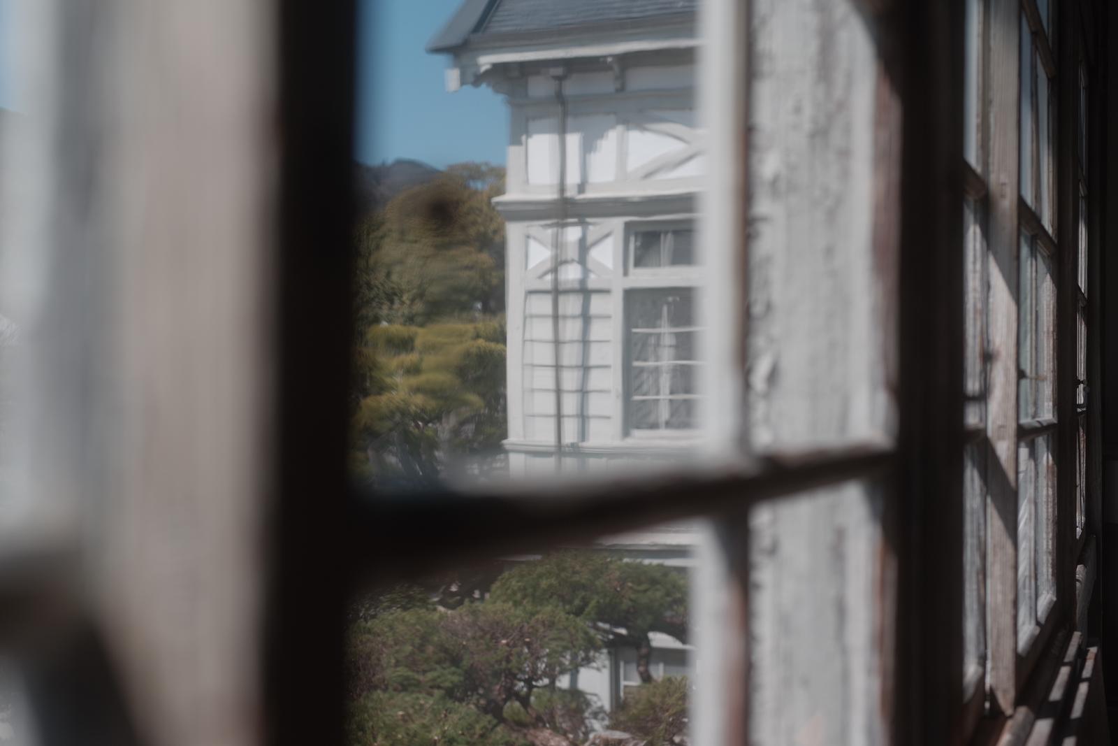 窓ガラス越しに見た旧遷喬尋常小学校の白い校舎と周辺の樹木