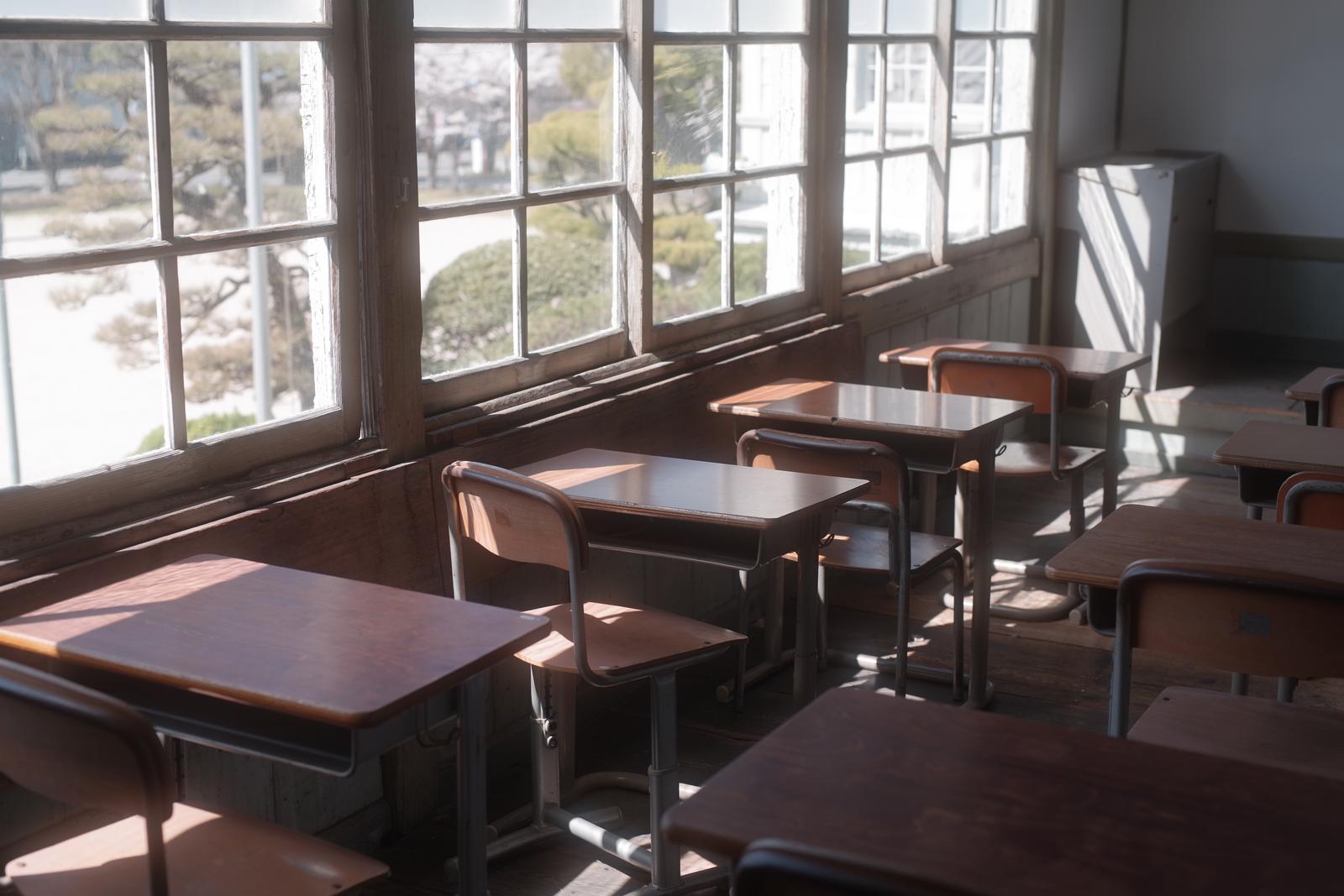 窓から日光が差し込む古い教室内に配置された木製の学習机と椅子