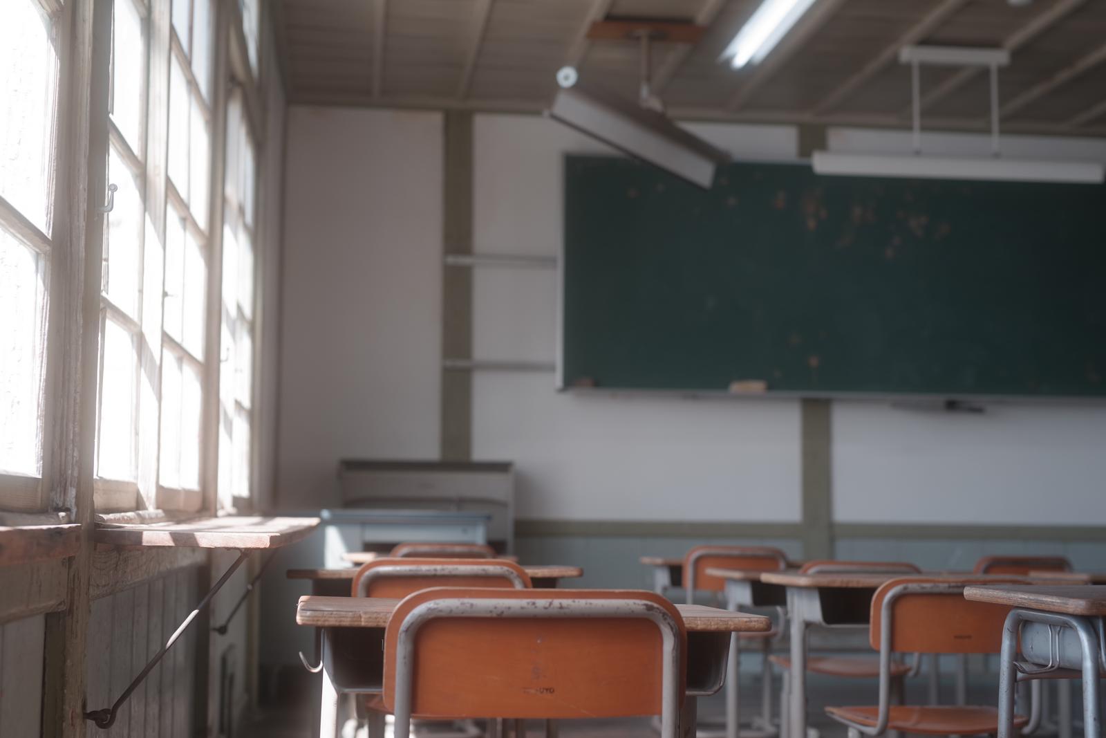 窓から光が差し込む放課後の教室。黒板と学習机、赤い椅子が並ぶ