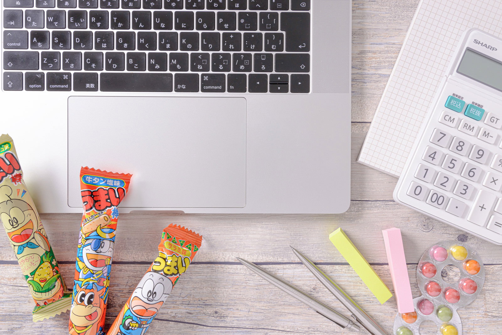 ノートパソコンと電卓、文房具、うまい棒などのお菓子が置かれたデスク