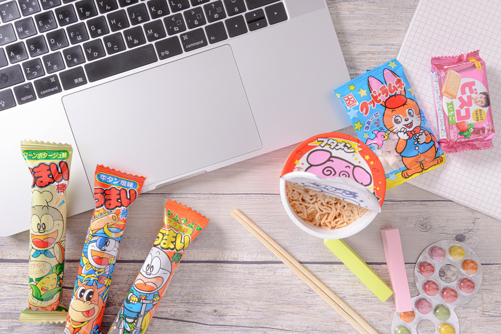 デスクのノートパソコン周りに並べられたうまい棒やブタメンなどのお菓子