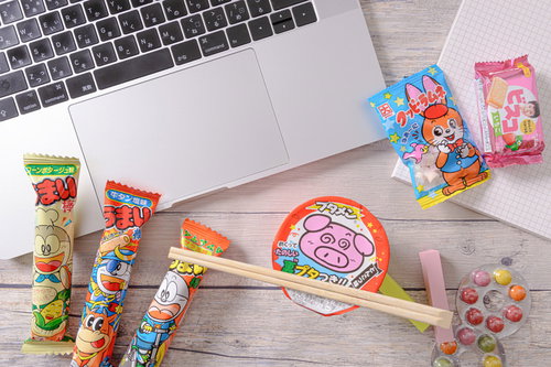 ブタメンやうまい棒に囲まれたノートパソコンのデスク風景
