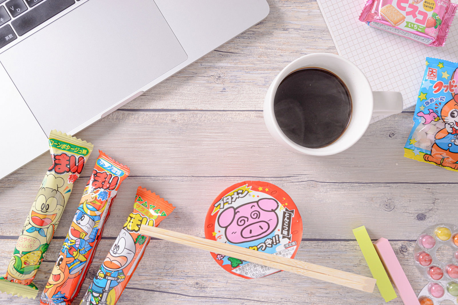 白い木目調のテーブルに置かれたコーヒーカップとブタメン、うまい棒などのお菓子類