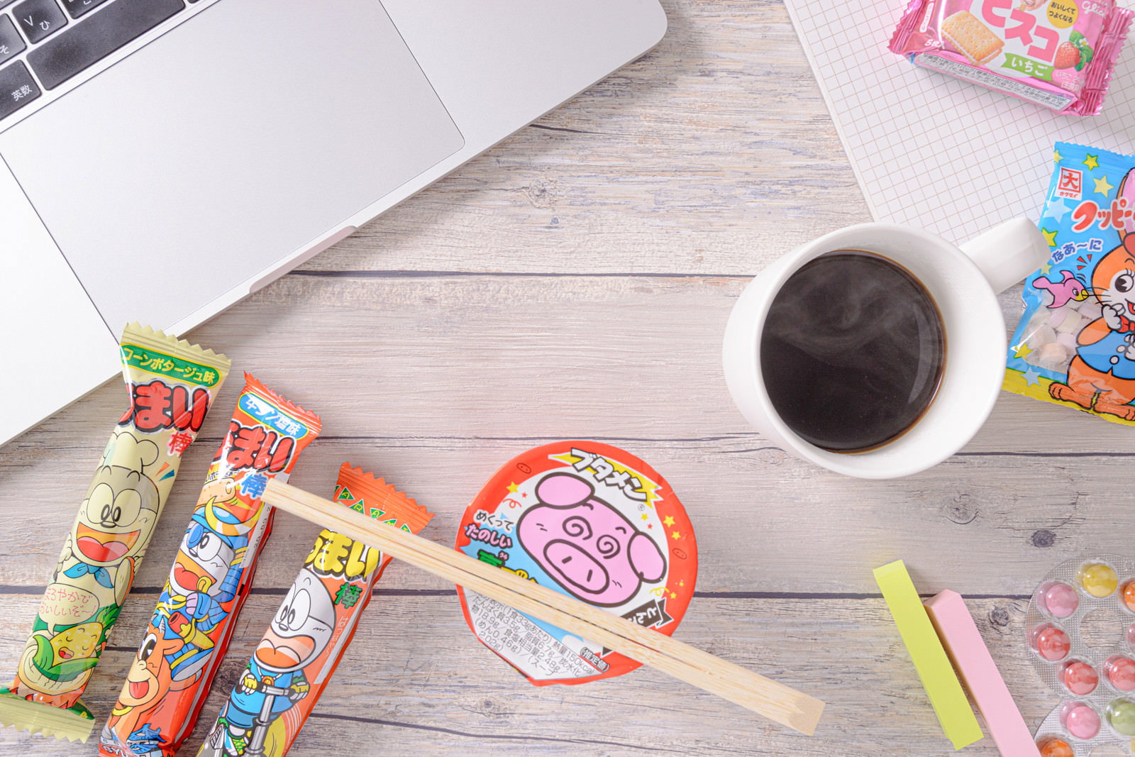 木のテーブル上のノートパソコン、ブタメンなどの駄菓子、コーヒーカップが配置された休憩風景