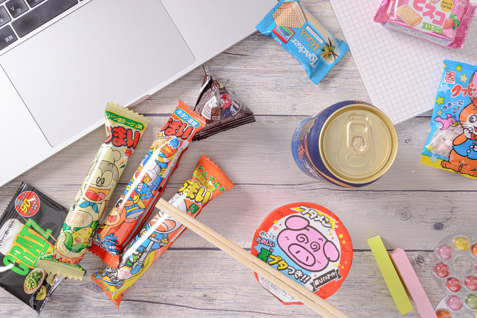デスク上のノートパソコンの横に置かれた缶ビールとうまい棒、ブタメンなどのお菓子