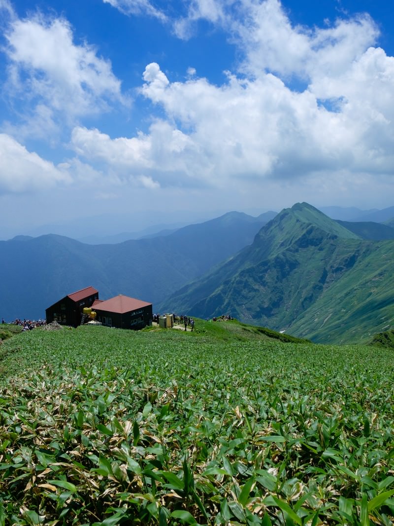青空の下、緑の山間に建つ赤い屋根の山小屋と背景の山峰