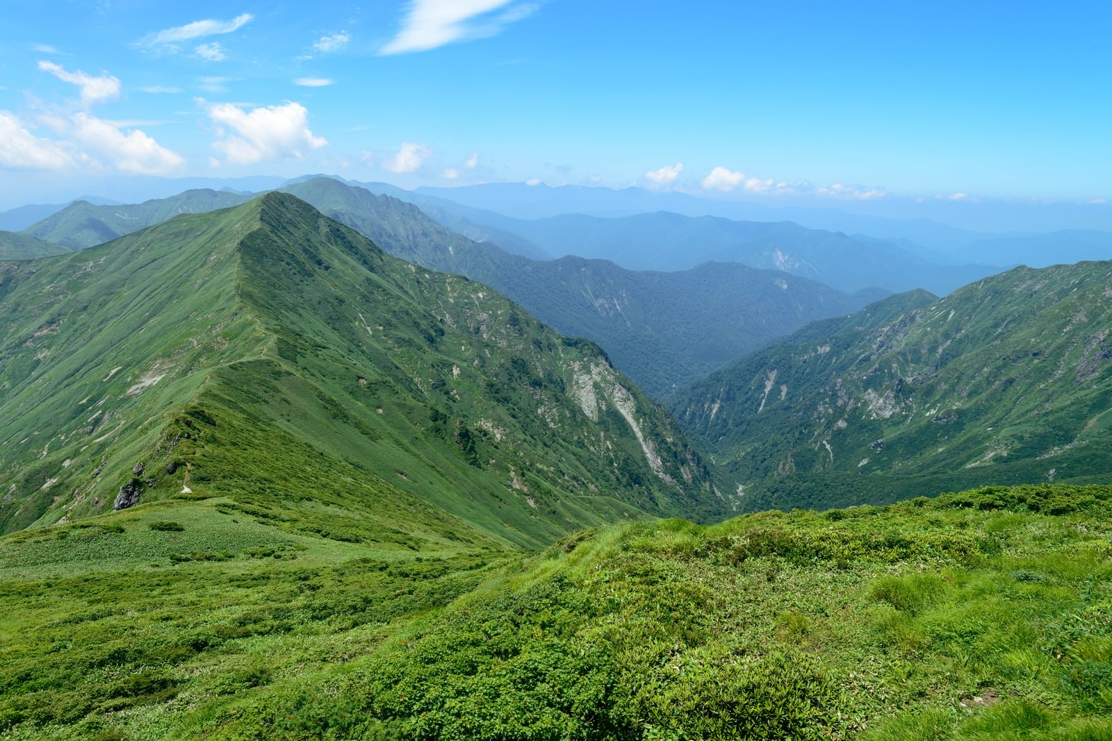 緑の山々が連なる稜線と渓谷、青空と雲が広がる山岳風景