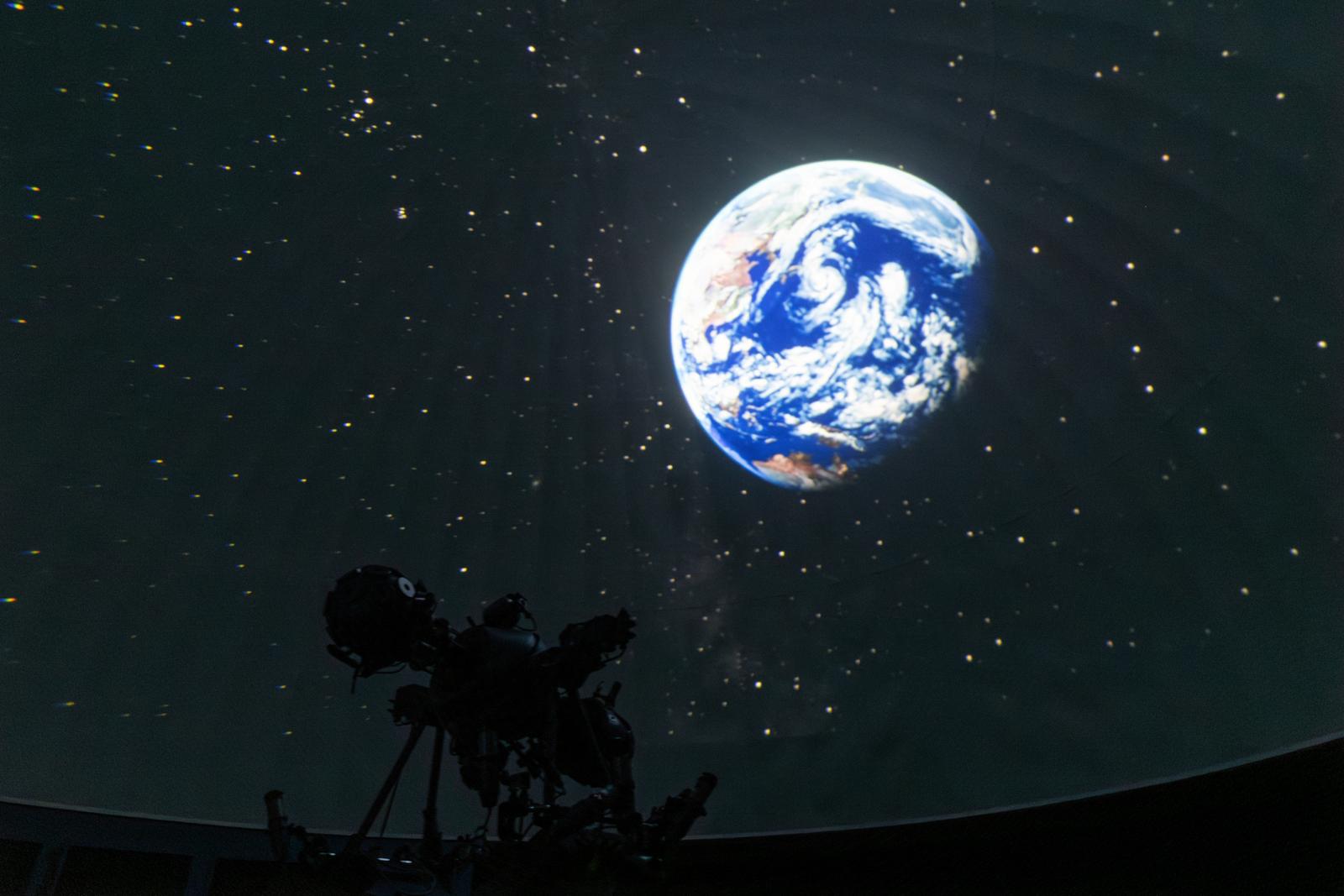 プラネタリウムに映し出される青白い地球と周囲の星空、望遠鏡のシルエット