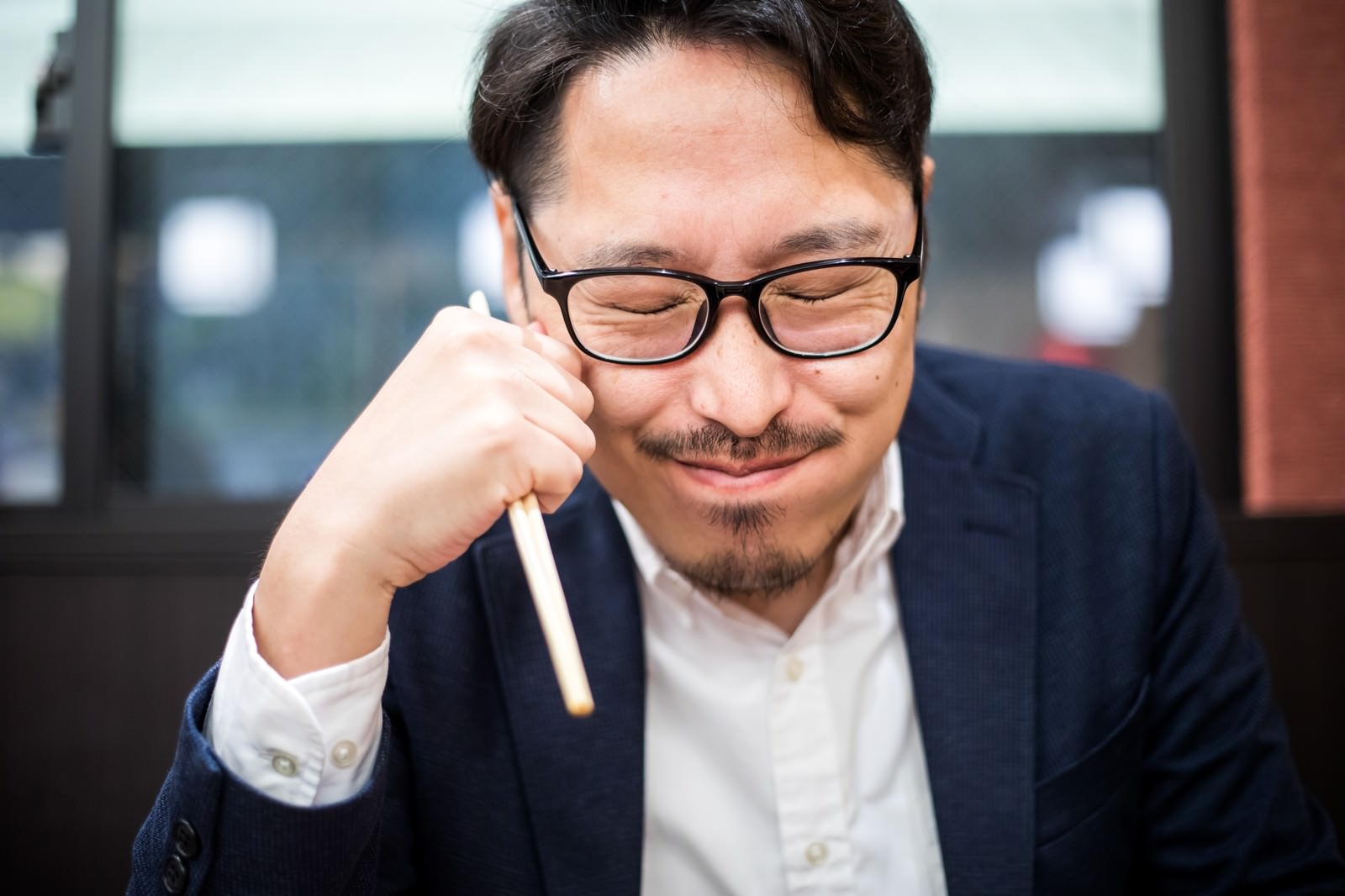 笑顔で頬杖をつく眼鏡をかけたスーツ姿の男性［モデル：ゆうせい］