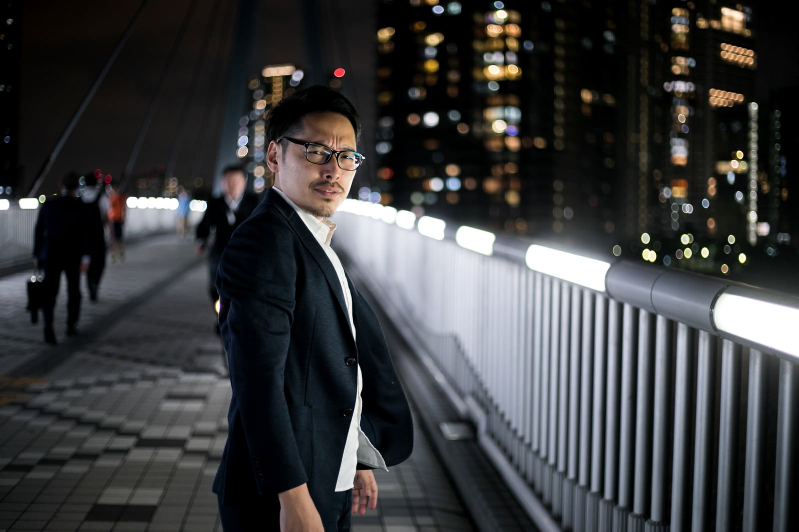 夜のビル街で立ち止まり、こちらを見つめるスーツ姿の眼鏡をかけた男性［モデル：ゆうせい］