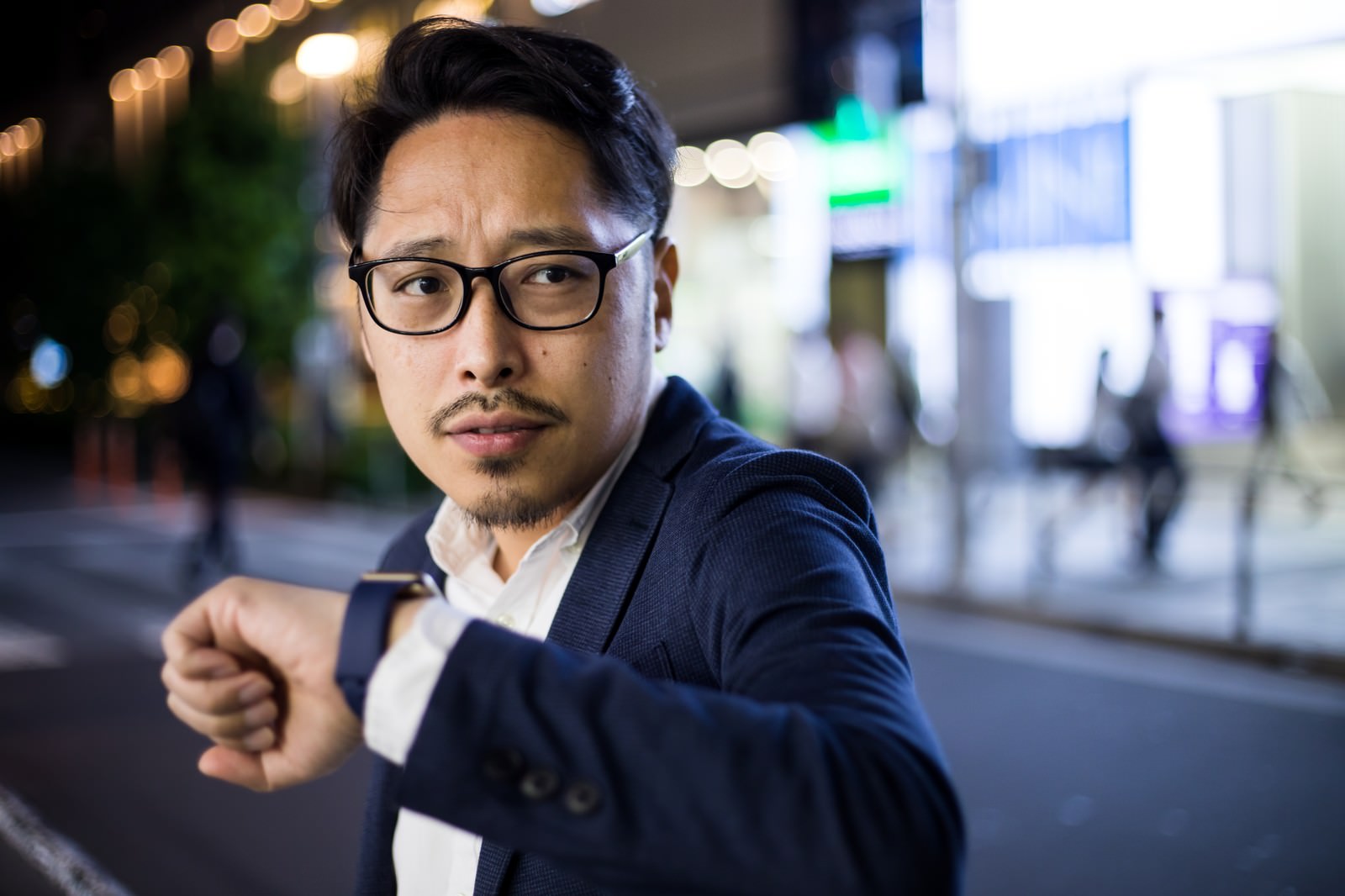 夜の街中で腕時計を見ながら待つスーツ姿の眼鏡をかけた男性［モデル：ゆうせい］