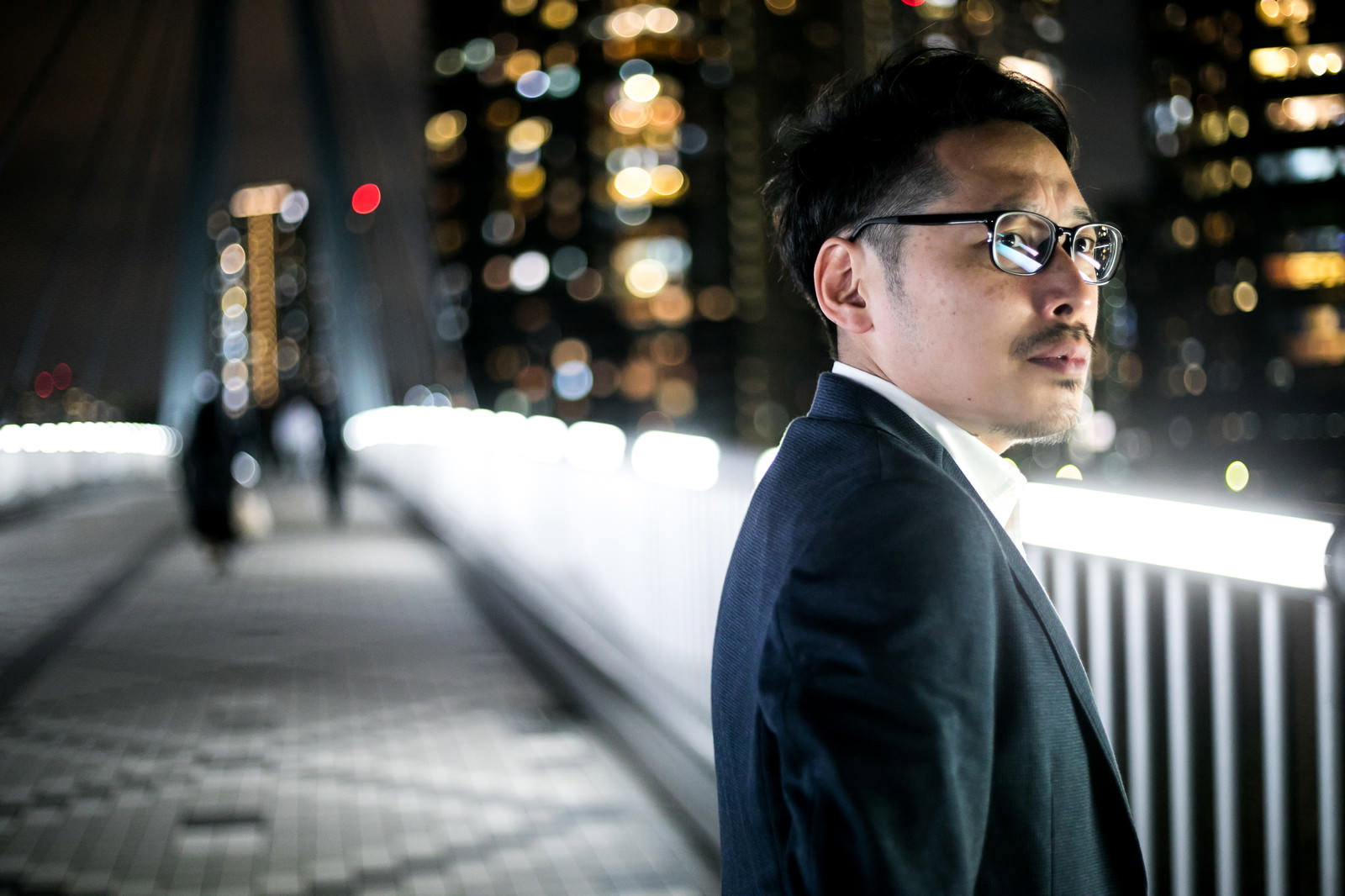 夜景を背景に立つ眼鏡をかけたスーツ姿の男性［モデル：ゆうせい］