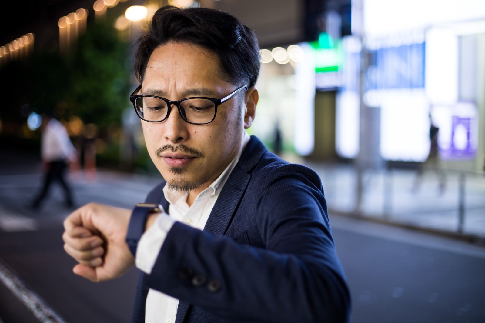 夜の街で時計を見つめるスーツ姿の眼鏡をかけた男性［モデル：ゆうせい］