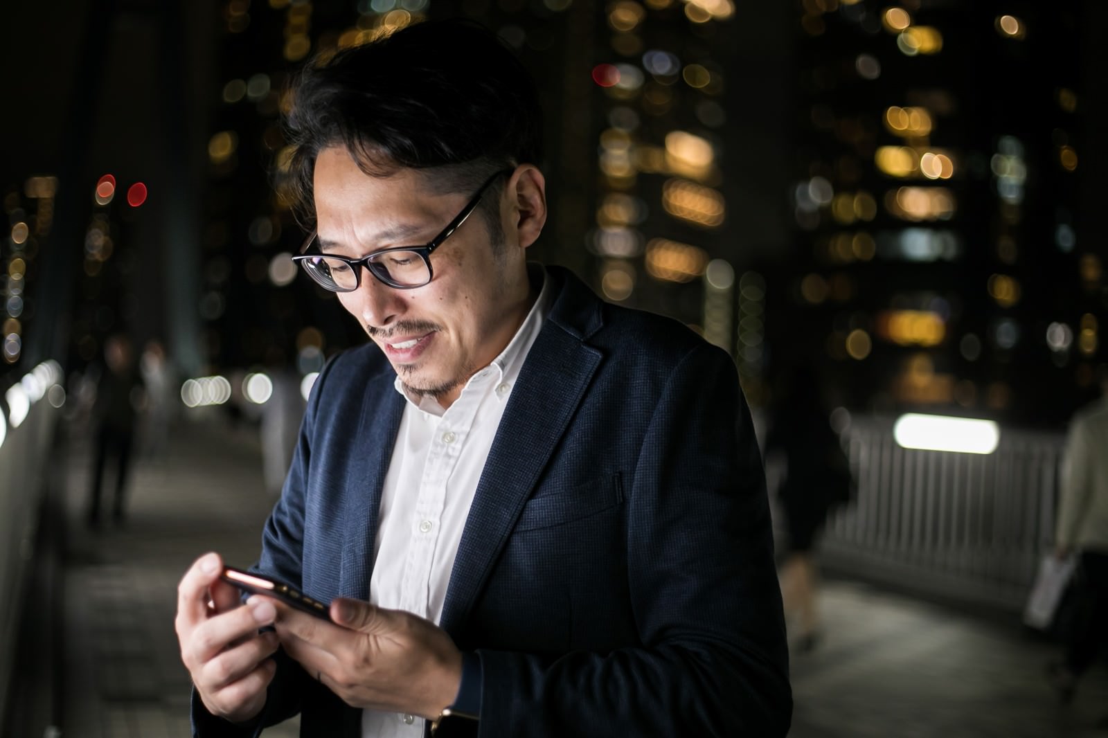 夜の街中でスマートフォンを見つめる眼鏡をかけた男性［モデル：ゆうせい］
