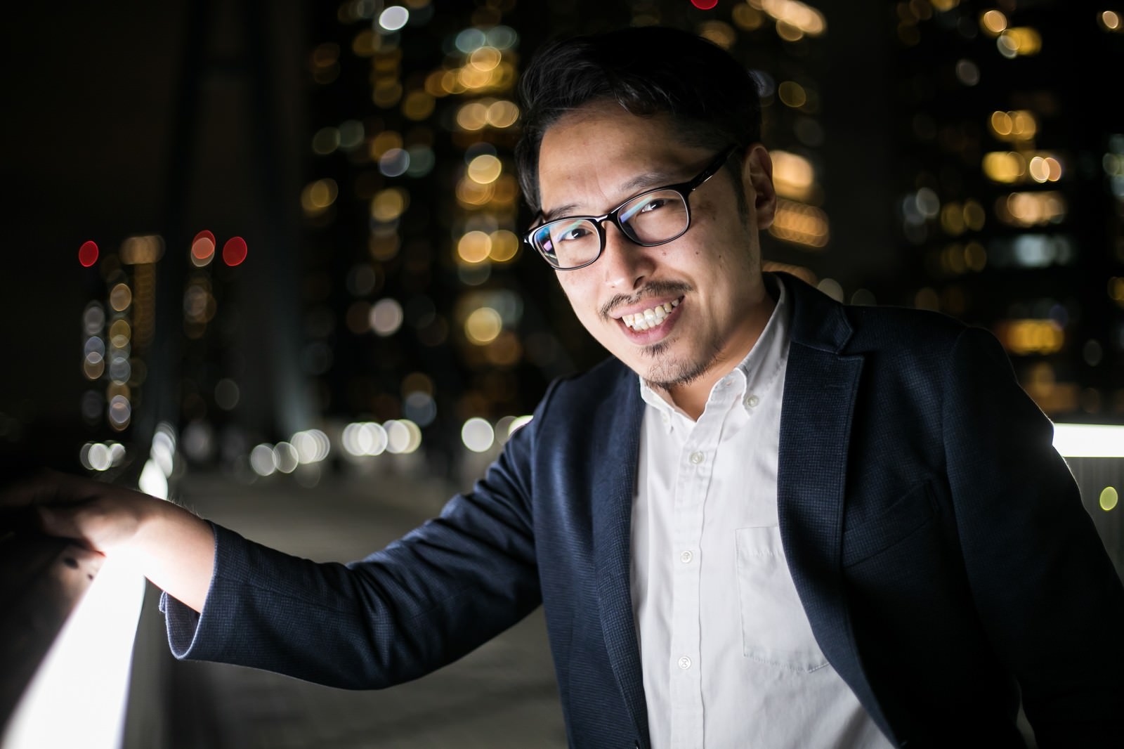 夜の街中でスマートフォンを見て笑顔を浮かべる眼鏡をかけたスーツ姿の男性［モデル：ゆうせい］