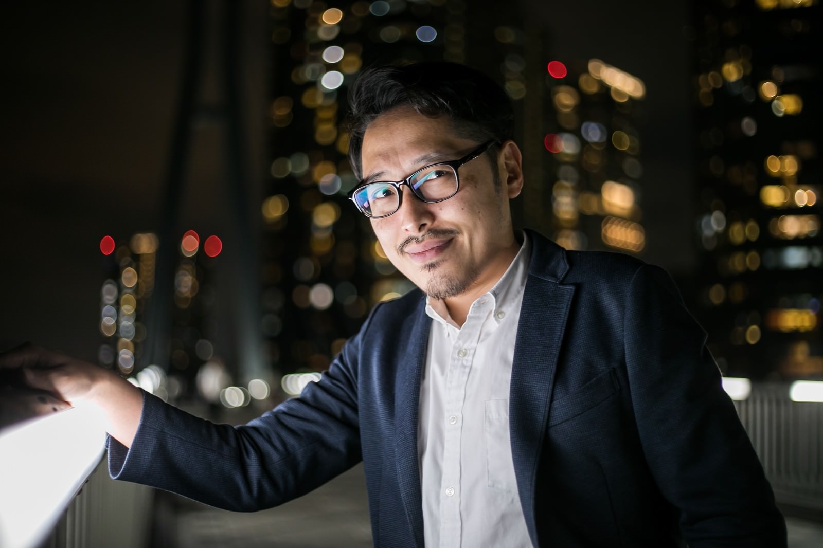 夜景をバックに立つスーツ姿の眼鏡をかけた男性［モデル：ゆうせい］