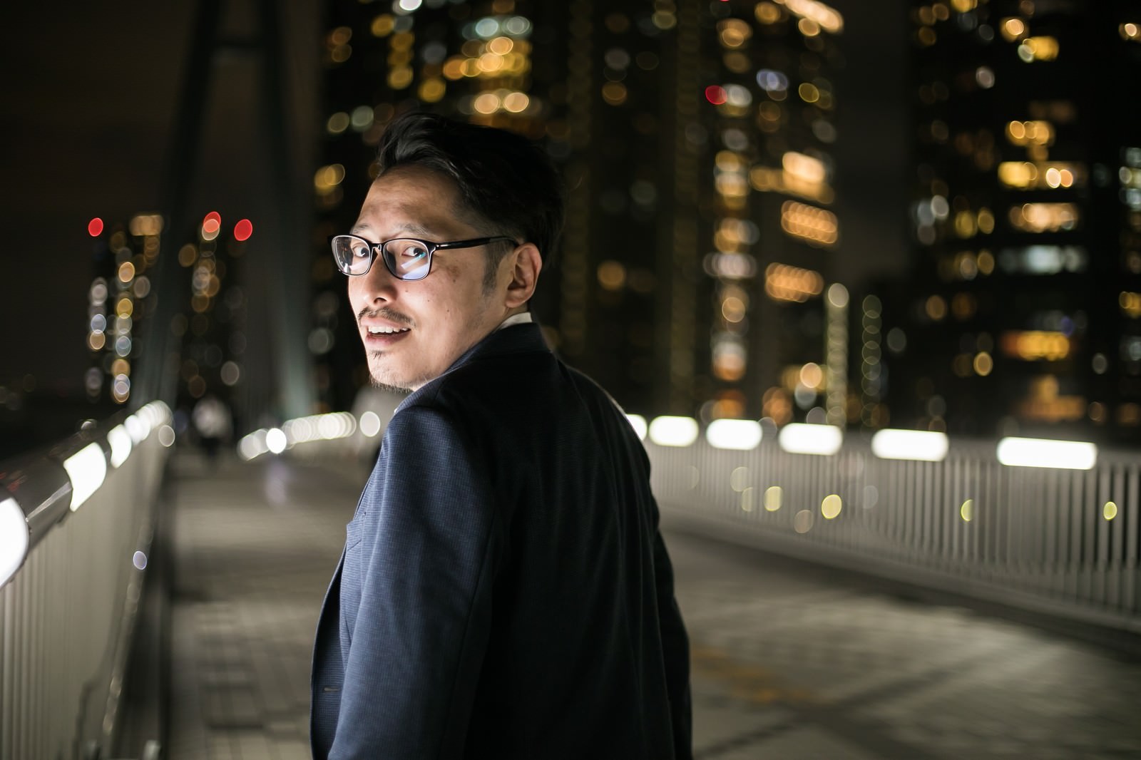 夜の道路で街灯に照らされながら振り返る眼鏡をかけた男性［モデル：ゆうせい］