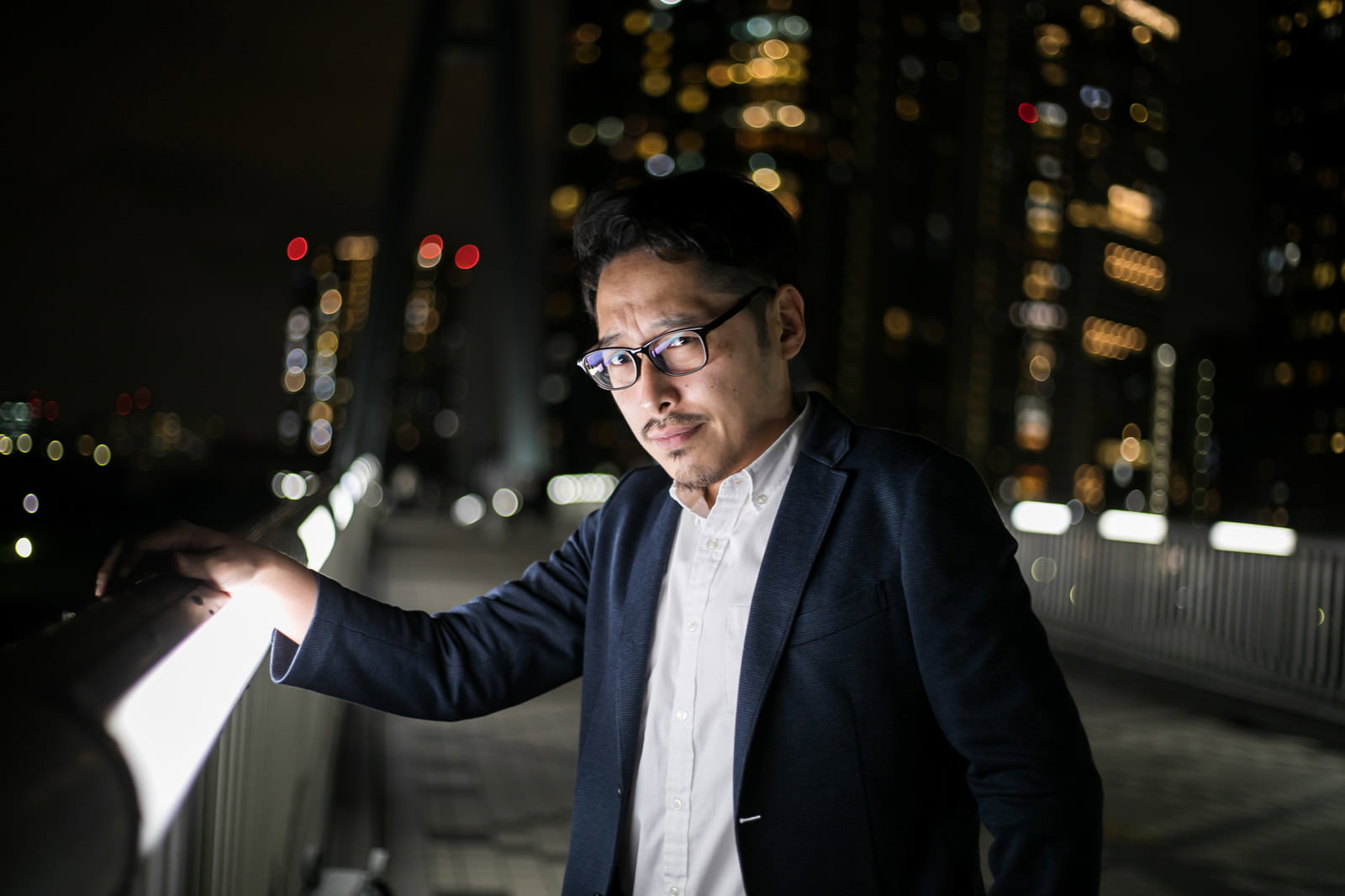 夜の街並みを背景に、スーツと眼鏡姿で真剣な表情を浮かべる男性が手すりに肘をかけている［モデル：ゆうせい］